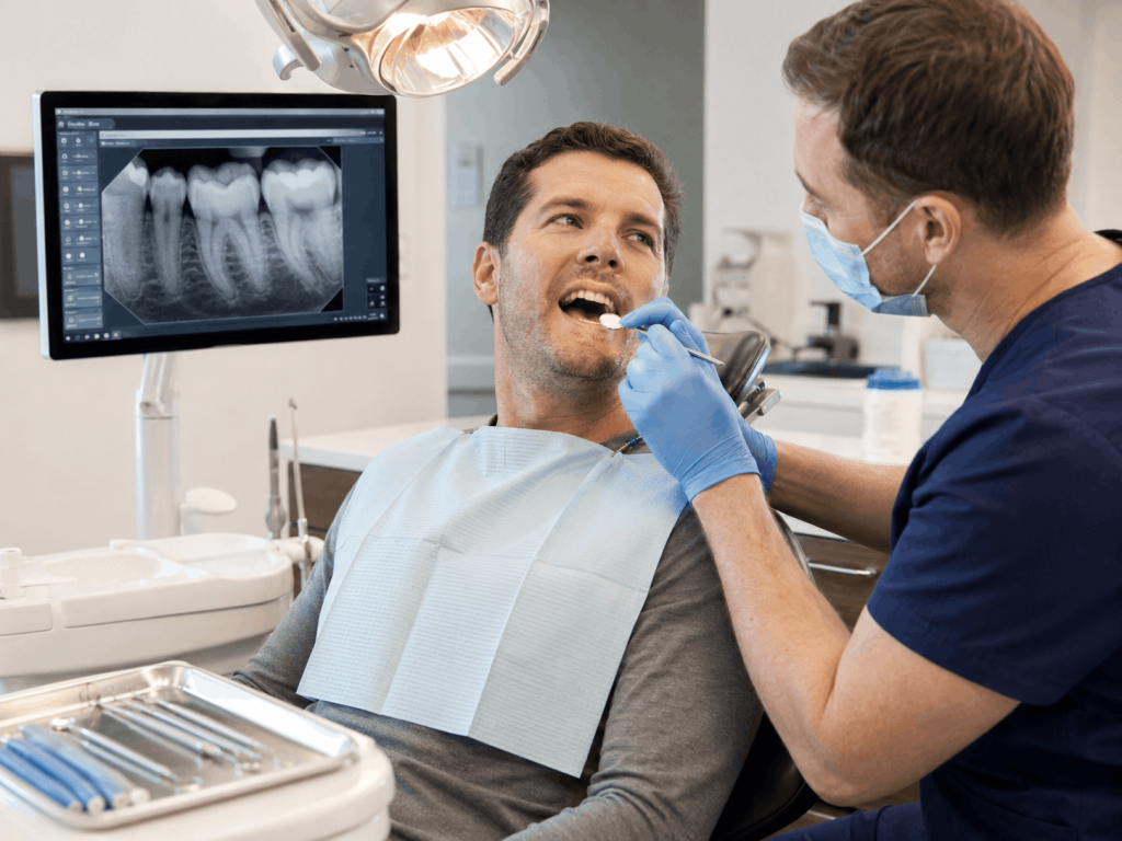An adult patient sits in a bright emergency dental clinic while a dentist examines a cracked back tooth using a mirror and focused exam light. A dental X-ray is displayed on a nearby monitor, and the patient’s tense expression begins to ease as the dentist calmly explains the damage and possible treatment options.
